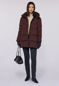 Vinröd vadderad jacka med huva, med dragkedja och tryckknappstängning, bärs över en beige tröja, kombinerad med svarta leggings och handskar.