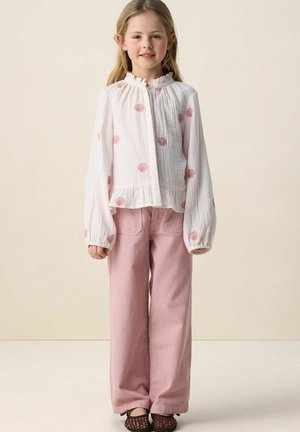 Jeune fille aux longs cheveux blonds portant un chemisier blanc orné de coquillages roses, un pantalon en velours côtelé rose et des chaussures tissées marron, debout devant un mur neutre.