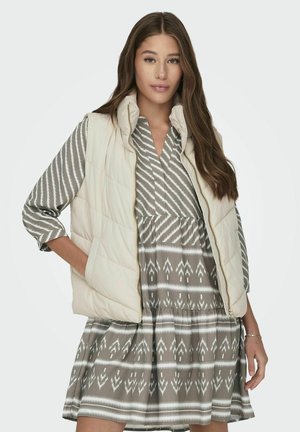 Gilet matelassé beige avec un col haut porté sur une robe grise à motifs, comportant des rayures horizontales et verticales. Fermeture éclair et poches latérales.