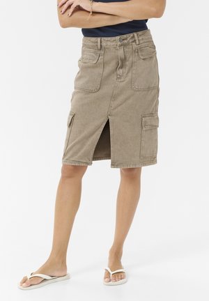 Jupe cargo beige longueur genou avec fente avant et multiples poches, portée avec des tongs blanches et un haut sombre.