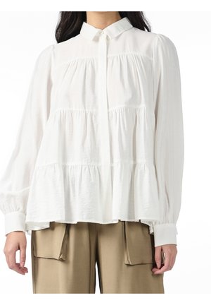 Femme portant un chemisier blanc à manches longues avec volants et poignets boutonnés, ainsi qu'un pantalon beige avec de grandes poches avant.