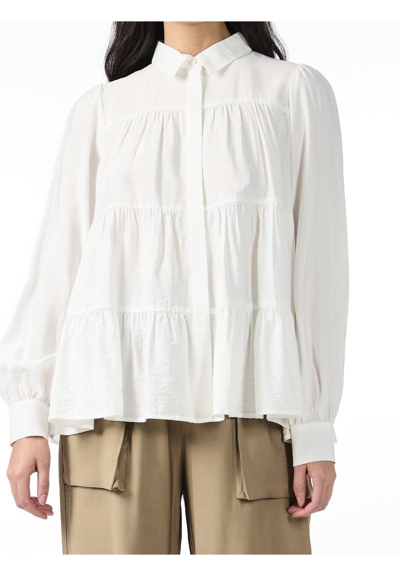 Femme portant un chemisier blanc à manches longues avec volants et poignets boutonnés, ainsi qu'un pantalon beige avec de grandes poches avant.