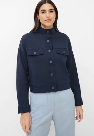 Femme portant une veste boutonnée bleu marine avec des poches poitrine à rabat et un pantalon taille haute bleu clair, debout devant un fond uni.