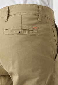 Pantalones caqui con una textura suave, que presentan un bolsillo trasero, costuras sutiles y un pequeño logo naranja de "BOSS" en la línea del cinturón.