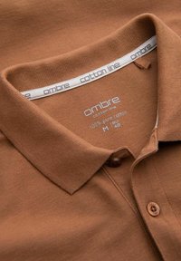 Ruskea puuvillapolo-paita, jossa on teksturoitu kangas, kolme napinläpeä ja kauluksessa etiketti, jossa lukee "ombre cotton line".