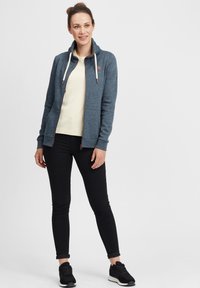 Blå zip-huvtröja med snodd, bärs över en ljusgul skjorta, kombinerad med svarta slim-fit byxor och svarta sneakers.