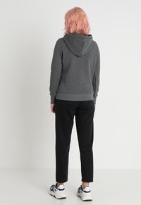 Persona con capelli rosa che indossa una felpa grigia scura, pantaloni neri e sneakers bianche, in piedi di fronte a uno sfondo bianco semplice.