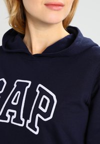Kvinna som bär en mörkblå hoodie med en stor vitomgärdad textlogotyp på bröstet, visar endast läppar och nedre ansikte.