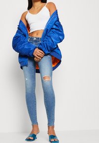 Kvinna i en vit croptop, en stor blå jacka med orange foder, rivna skinny jeans och blå klacksandaler, står med händerna knäppta.