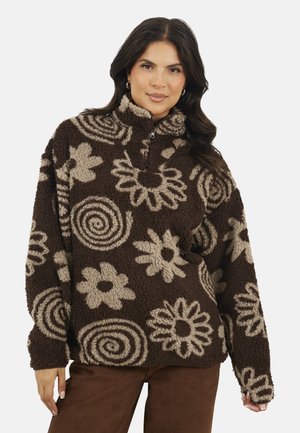 Vrouw in een bruin en beige geblokt fleece sweater met een halve rits en bloem- en spiraalontwerpen, gecombineerd met een paarse broek.