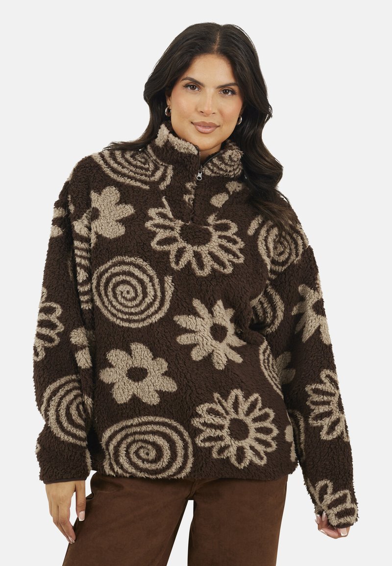 Donna che indossa un pullover in pile marrone e beige con un mezzo zip e disegni floreali e a spirale, abbinato a pantaloni marroni.