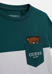Teal og hvid T-shirt med kort ærme og en bjørnebroderi på brystlommen. Har en rund hals og en blød stofstruktur.