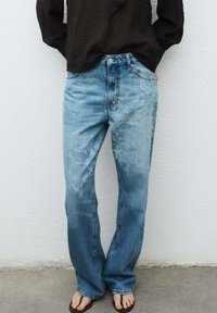 Jeans de denim azul claro con un corte relajado, detalles deslavados y diseño de cinco bolsillos. Combinados con una camiseta negra de manga larga y chanclas.