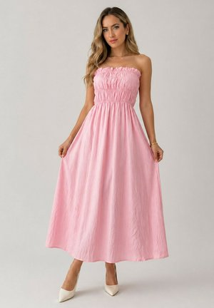 Femme portant une robe longue rose bustier smockée, tenant les côtés de la jupe, debout devant un fond clair uni avec des talons blancs pointus.