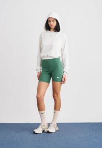 Bela jopa s kapuco in vrvico, zelene športne kratke hlače z logotipom Nike, bele poletne nogavice in lahki športni čevlji. Ozadje je enobarvno.