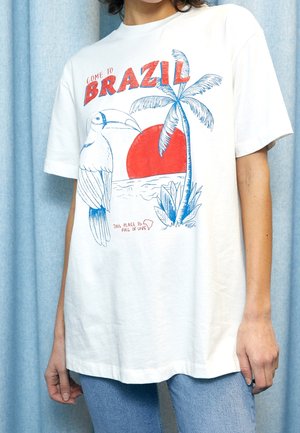Biały t-shirt z czerwono-niebieskim nadrukiem w stylu brazylijskim z tukaniem, palmą, czerwonym słońcem i tekstem "Przyjedź do Brazylii" oraz "To miejsce jest pełne miłości."