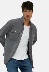 Graues Baumwollhemd mit zwei Vordertaschen, knöpfbarem Design und Kragen, über einem weißen T-Shirt getragen, kombiniert mit blauen Jeans.