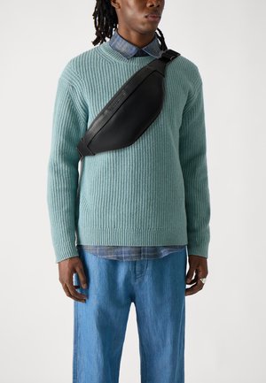 Een model draagt een gestructureerde mintgroene gebreide trui over een blauw geruit shirt en lichtblauwe spijkerbroek, met een zwarte crossbody tas.