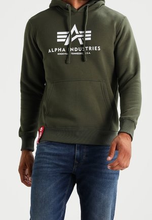 Man draagt een olijfgroene Alpha Industries hoodie en blauwe spijkerbroek, staand met ontspannen armen tegen een effen achtergrond.