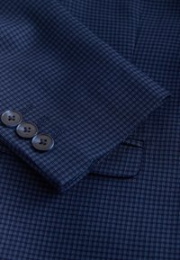 Primer plano de la manga de un traje azul marino a cuadros con tres botones oscuros y parte de un bolsillo a juego del saco visible.