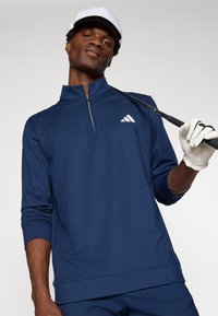 Námořnický golfový pulovr s půlzipem, dlouhými rukávy a bílým logem na hrudi. Hladký povrch, lehká látka, v kombinaci s bílou čepicí a rukavicemi.