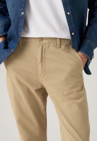 Beige katoenen broeken met een rechte pasvorm, voorzien van een knoopsluiting, riemlussen en zijzakken. Gaat gepaard met een denim overhemd.