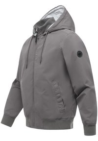 Veste à capuche grise avec fermeture éclair avant, poignets et ourlet côtelés, poches latérales, et un patch logo circulaire sur la partie supérieure de la manche.