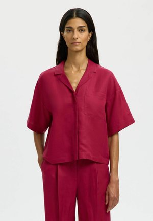 Femme aux longs cheveux bruns portant une chemise rouge à manches courtes et ample avec une poche sur la poitrine, accompagnée d'un pantalon rouge assorti, sur un fond uni.