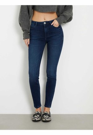 Mujer con jeans ajustados azul oscuro, suéter gris corto que deja ver el abdomen y zapatos sin cordones con estampado, de pie frente a una pared lisa.