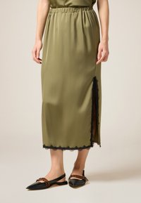 Gonna midi verde oliva con vita elastica, caratterizzata da un orlo in pizzo nero e uno spacco laterale. Abbinata a ballerine nere a punta e dettagli a righe.