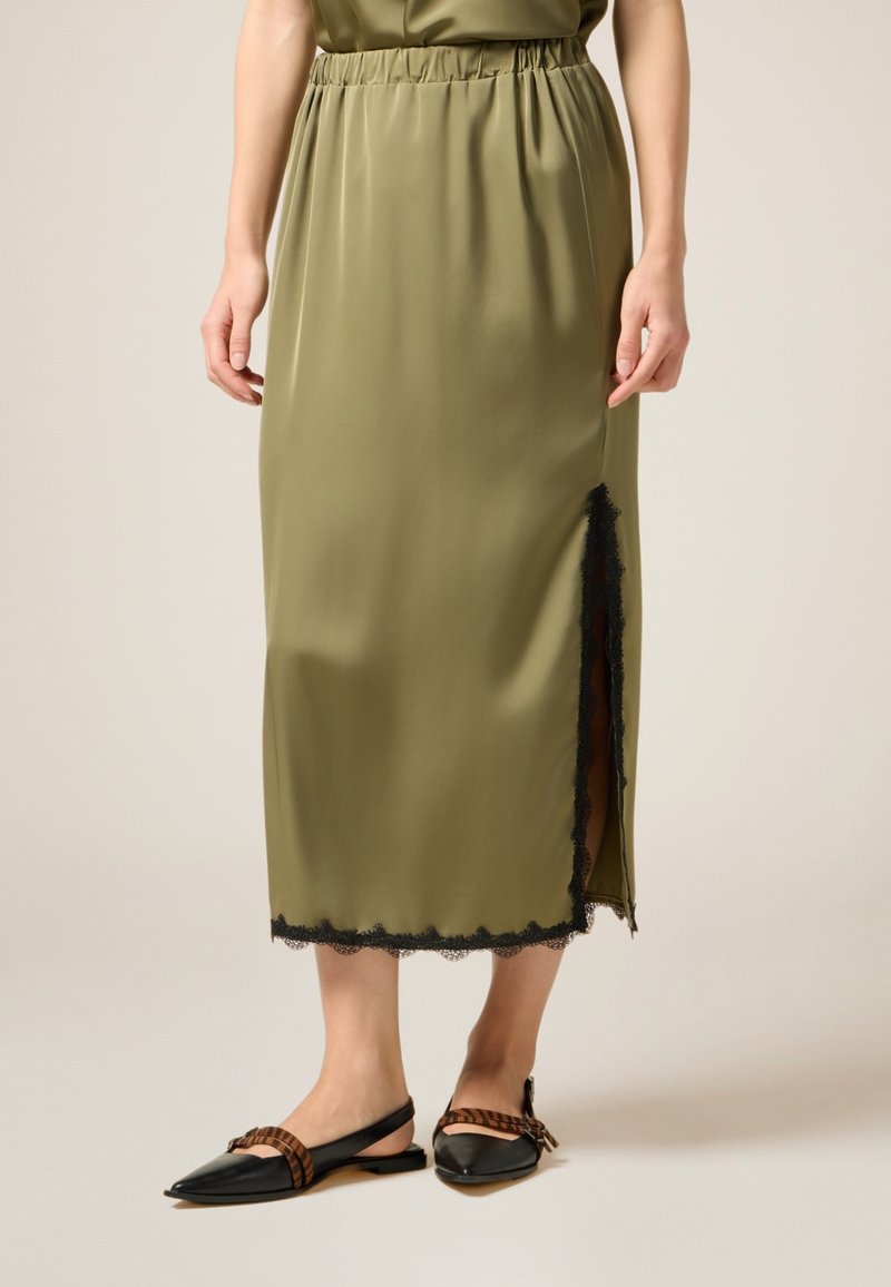 Gonna midi verde oliva con vita elastica, caratterizzata da un orlo in pizzo nero e uno spacco laterale. Abbinata a ballerine nere a punta e dettagli a righe.