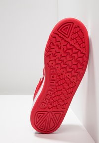Červená gumová podešev boty s texturovanými vzory gripu, včetně herringbone designu a brandingu. Boční strana obsahuje bílé akcenty a švy.