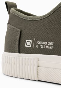 Grüne Leinensneaker mit einer weißen Gummisohle, die eine strukturierte Seite aufweist, und dem Text "DEIN EINZIGES LIMIT IST DEINER GEIST" in Weiß bedruckt.
