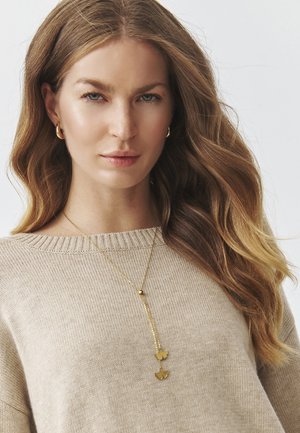 Gul vedhæng necklace med to blomst-formede charms, båret over en lyse beige, ribbet sweater. Lang, tynd kæde med en kugle accent.