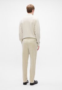 Calças de sarja bege com dois bolsos traseiros, combinadas com um sweatshirt bege claro e sapatos pretos, vistas de trás contra um fundo branco.