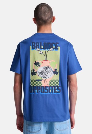 Mężczyzna w niebieskim t-shircie z grafiką różowego wazonu z kwiatami, czarnych ptaków, zielonej szachownicy i napisem „Balance Opposites” na plecach.