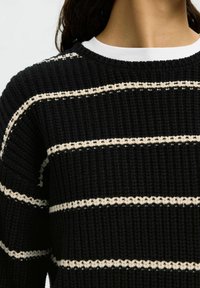 Maglione nero lavorato a maglia con strisce orizzontali color crema, caratterizzato da un collo rotondo e un tessuto testurizzato. Vista ravvicinata della parte superiore.