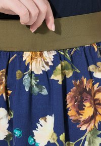 Kukallinen hame tummansinisellä taustalla, jossa on oransseja ja vaaleanbeigejä kukkia, sekä sileä oliivivihreä joustava vyötärö.