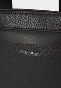 Černá texturovaná syntetická taška s hladkým povrchem, s elegantním designem a stříbrným logem "Calvin Klein". Minimalistická a odolná.