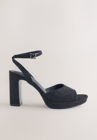 Sort ruskind højhælet sandal med bred åben tå, en tyk blokhæl og en justerbar ankelrem med en metal spænde.