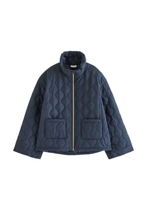 Ung kvinde står iført en mørkeblå quiltet jakke, falmede blå jeans, burgunder sko og en lilla top mod en ensfarvet lys baggrund.