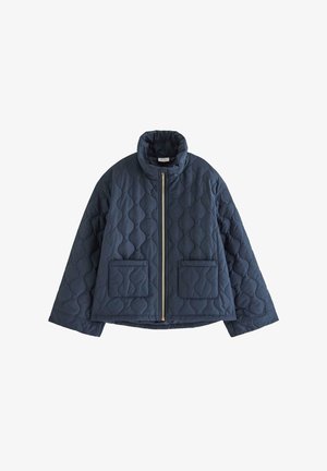 Jeune femme debout portant une veste matelassée bleu marine, un jean bleu délavé, des chaussures bordeaux et un haut violet sur un fond clair uni.