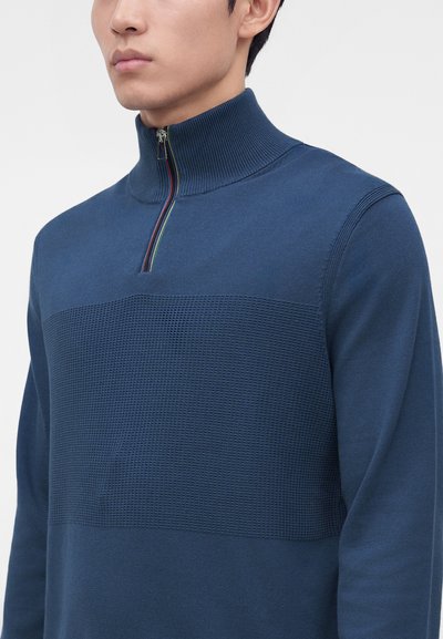 Homme portant un pull bleu texturé avec un col montant côtelé et une courte fermeture éclair avec des rayures colorées.