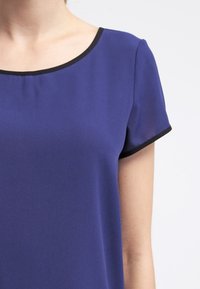 Fialová blůza s krátkými rukávy, černý lem kolem kulatého výstřihu, měkká textilie a čistý, minimalistický design.