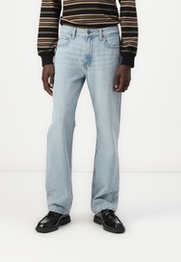 Jeans de mezclilla azul claro con corte recto, diseño de cinco bolsillos y un sutil efecto de desgaste. Llevados con una camisa negra a rayas y zapatos negros.