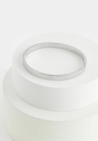 Sølv bangle-armbånd med en glat, poleret overflade og en let åbning; cirkulær form hviler på en hvid, tekstureret display.