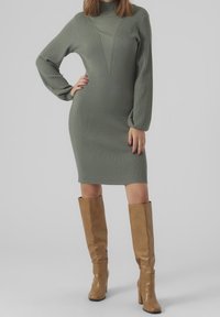 Robe côtelée à manches longues, longueur genou, de couleur vert sauge, avec un col montant et un décolleté en V. Associée à des bottes hautes couleur beige.