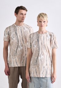 To personer har på seg kortermede, lyse beige topper med et abstrakt mønster i blå og kremfarger, med rund hals.