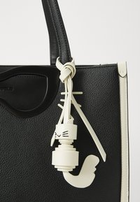Crna teksturirana torba s kontrastnim bijelim privjeskom u obliku mačke, koja ima elegantnu naramenicu i sjajne metalne detalje.
