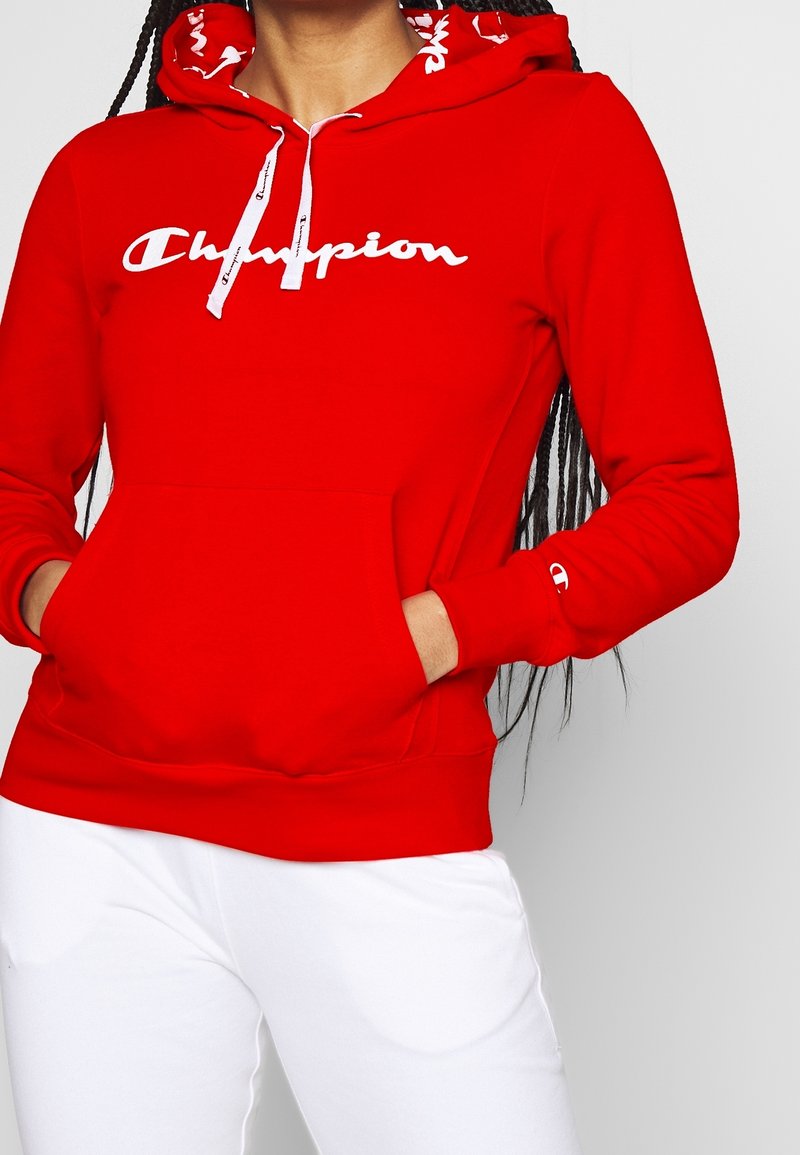 Personne portant un sweat à capuche rouge Champion avec des cordons blancs et un logo, mains dans la poche avant, associé à un pantalon blanc.